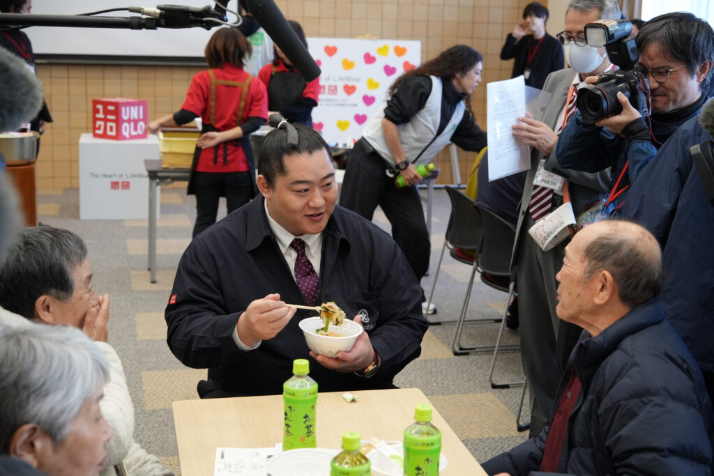 力士と町の人がちゃんこ鍋を食べている
